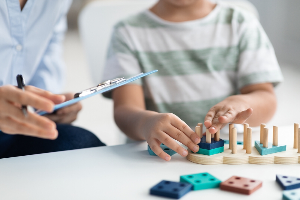 Little boy plays educational game during therapy session; specialist takes notes supporting future development planning.